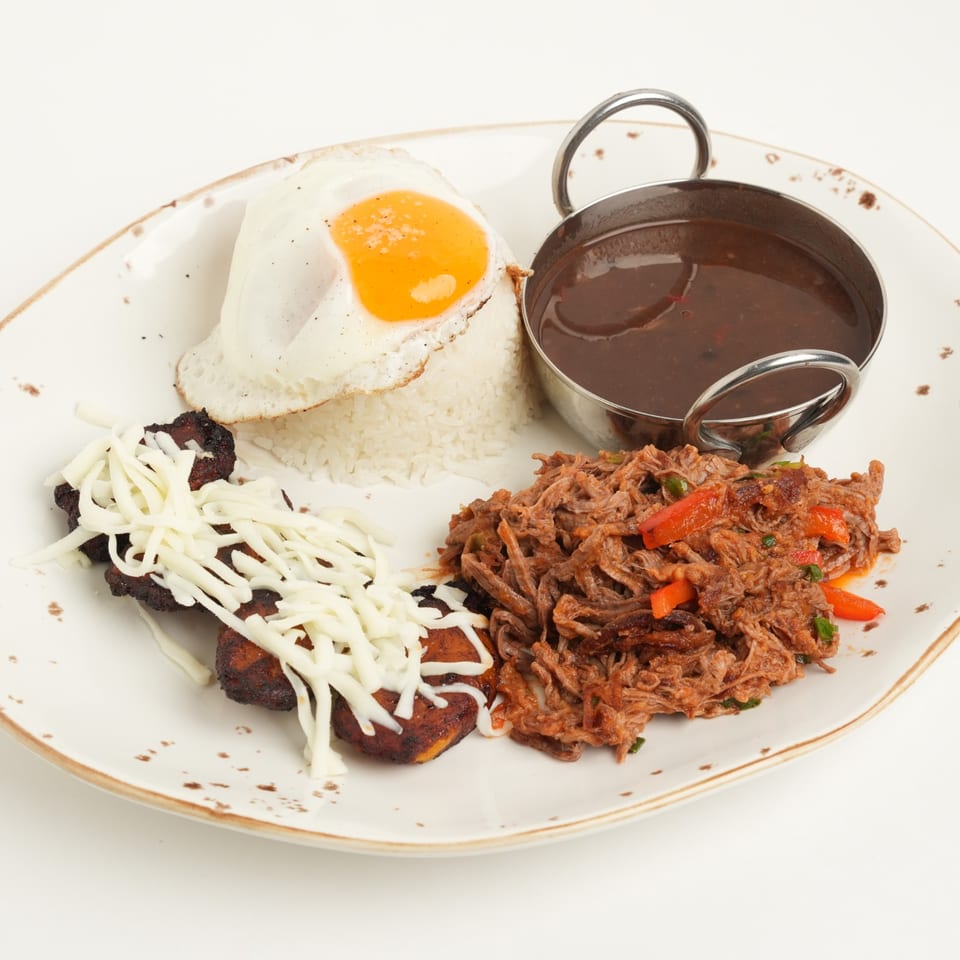 Plate of white rice topped with a fried egg, shredded beef, melted cheese, and a bowl of dark sauce.