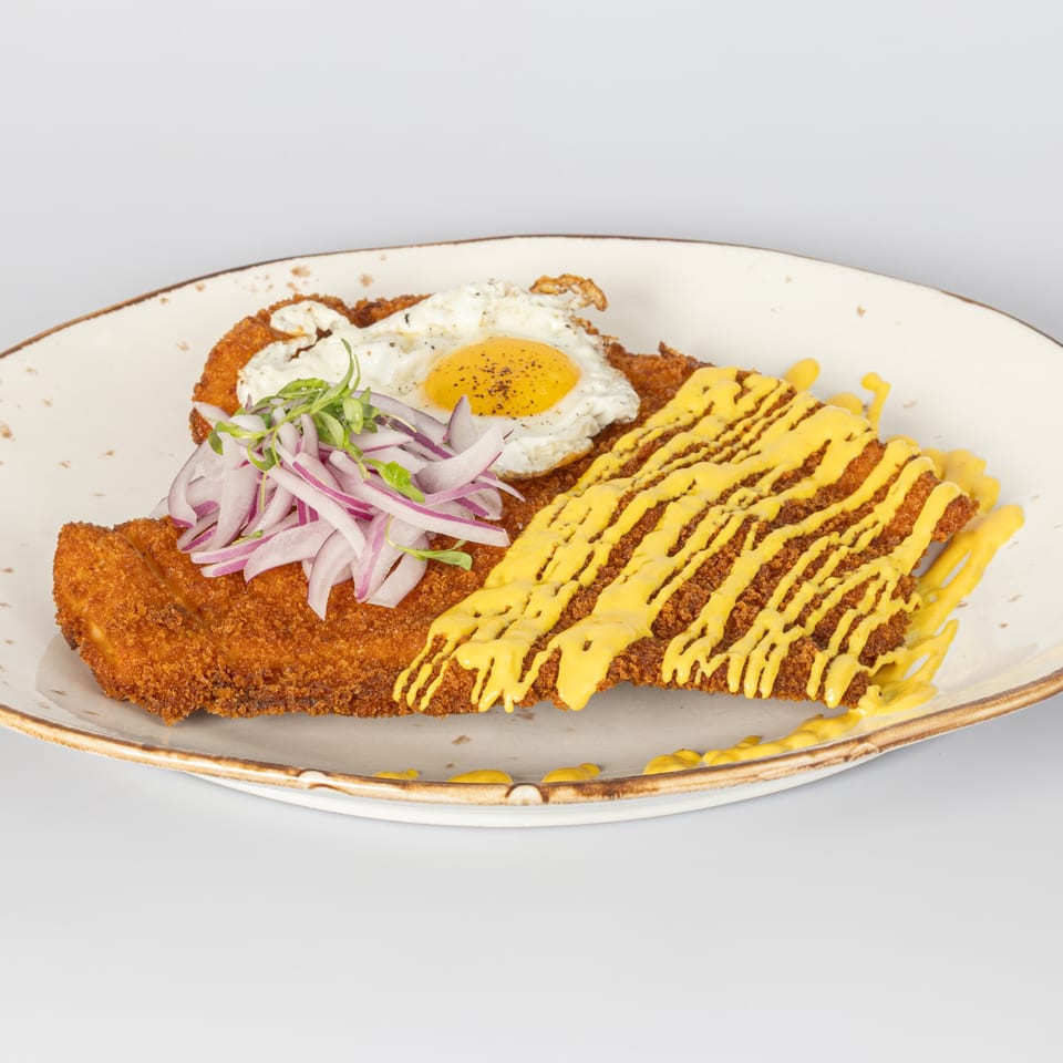 Two crispy fried chicken cutlets, one topped with a fried egg and red onions, the other with yellow sauce.