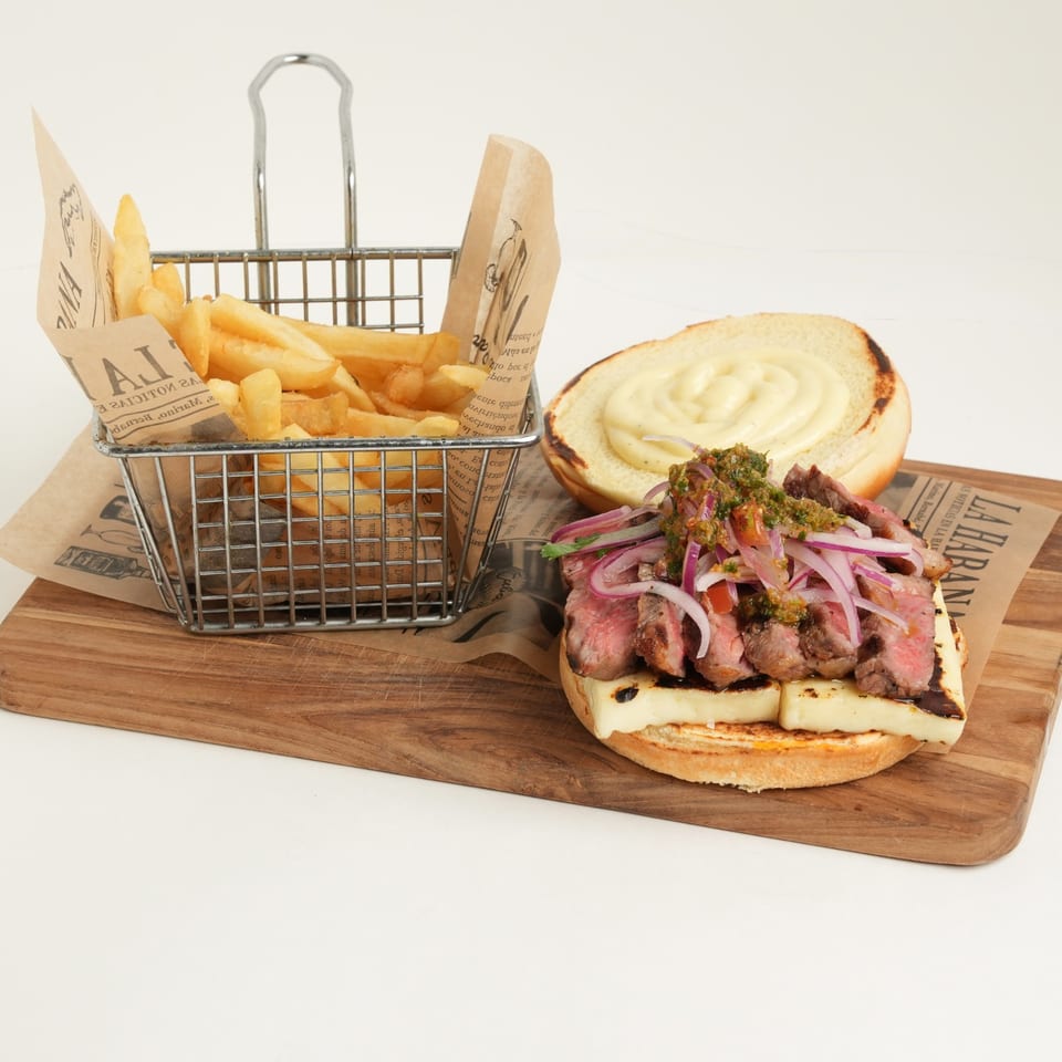 Steak sandwich with melted cheese, red onions, herbs, and sauce served beside fries in a wire basket on a wooden board.
