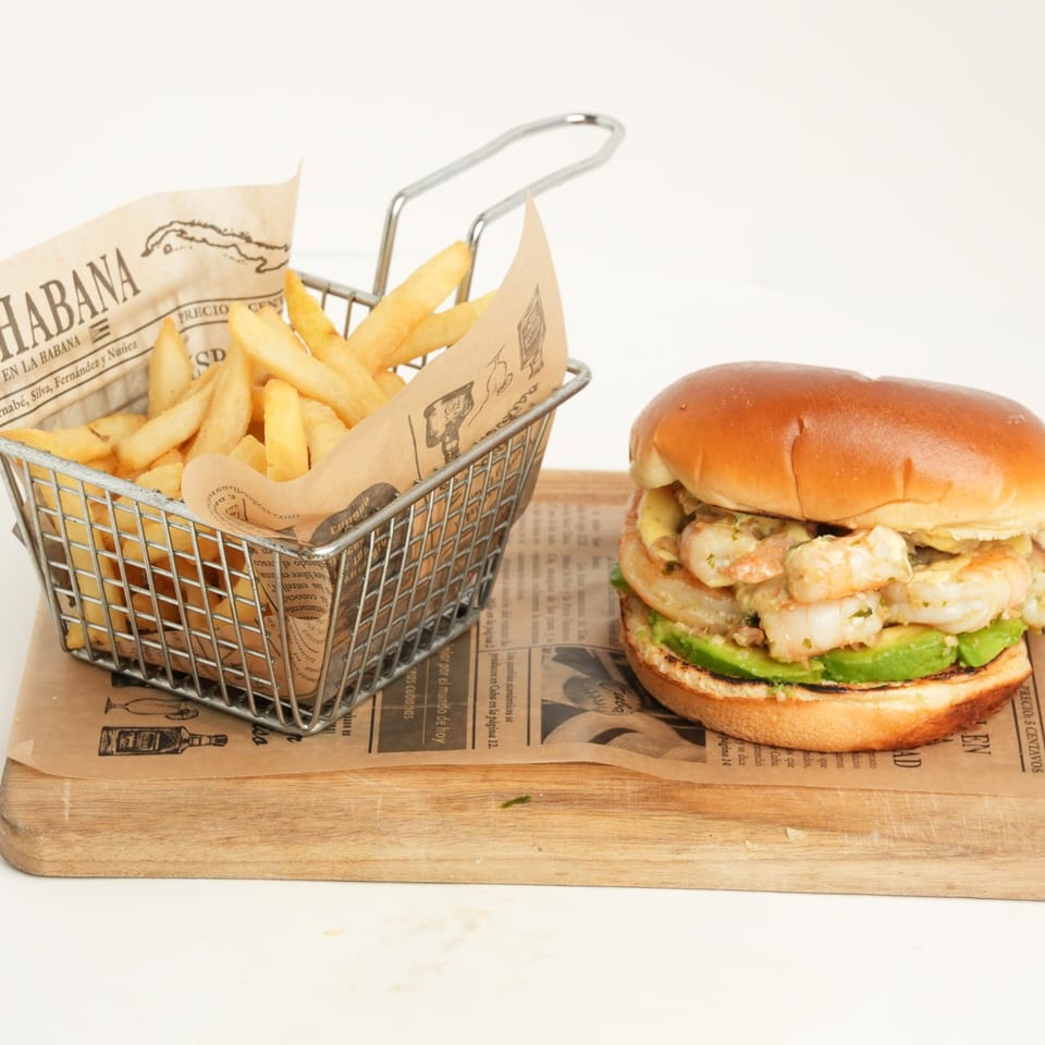Shrimp burger with avocado and fries in a metal basket on a wooden serving board.