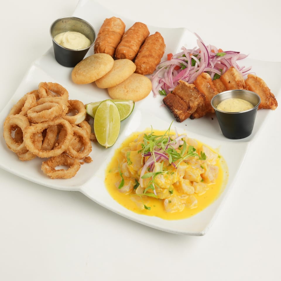 Large platter of fried seafood, golden croquettes, bread rolls, lime wedges, onion salad, creamy yellow ceviche, and dipping sauces.