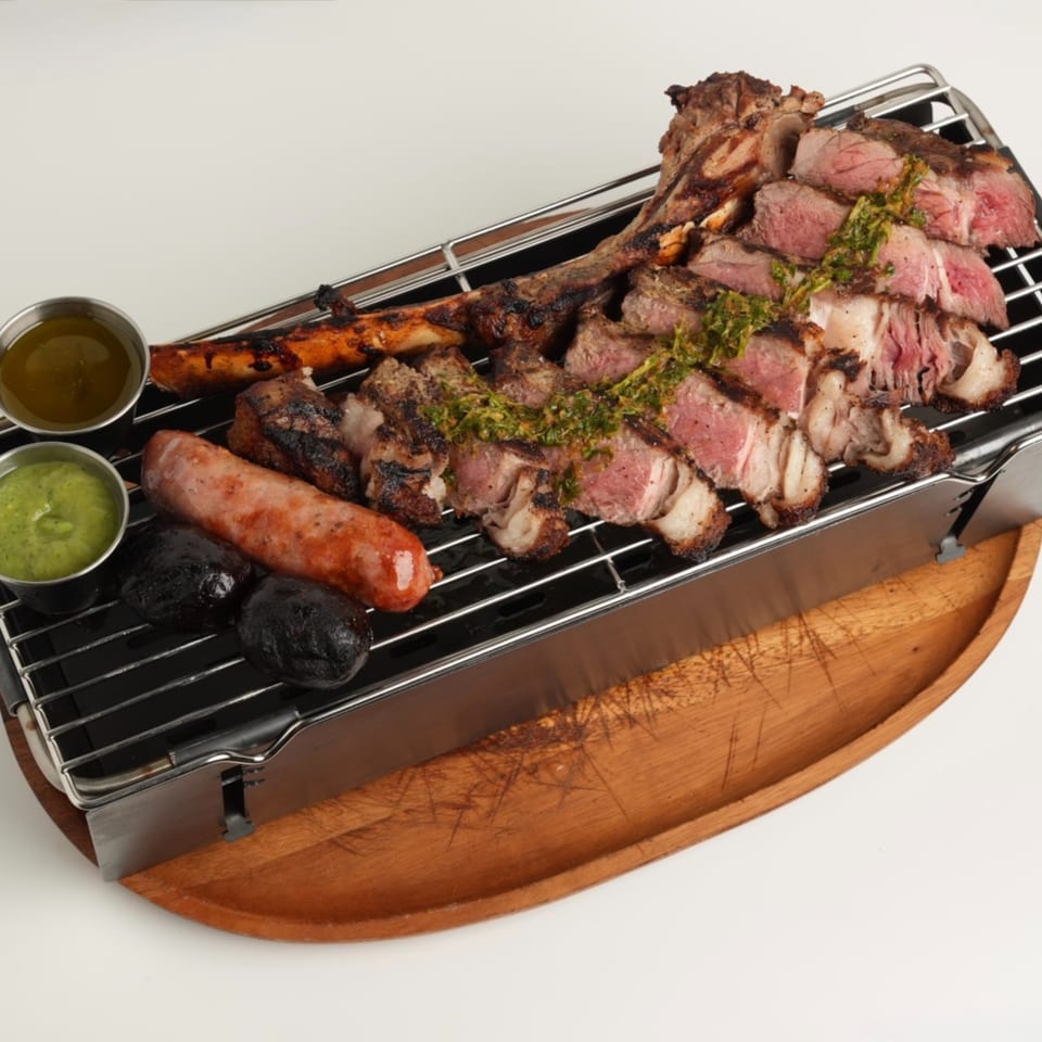 Grilled meats, sausages, and vegetables arranged on a tabletop grill with chimichurri and dipping sauces.