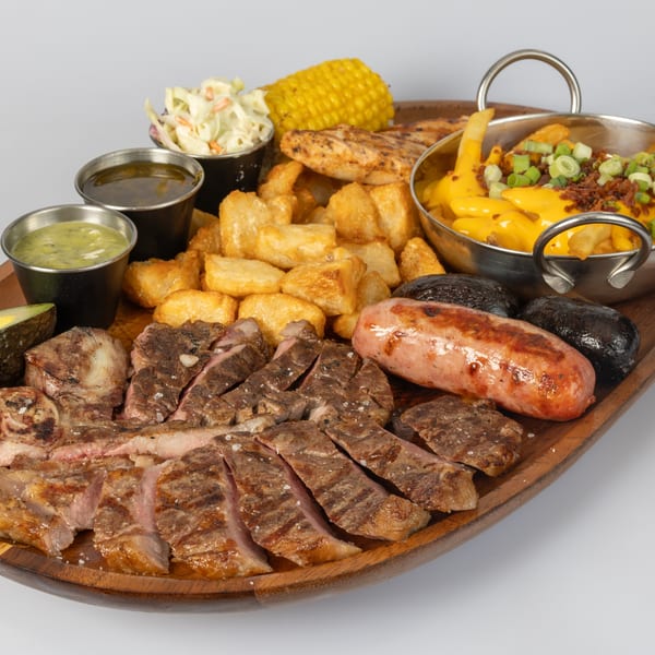 A large wooden platter with sliced grilled steak, sausage, fries, corn, coleslaw, and sauces.