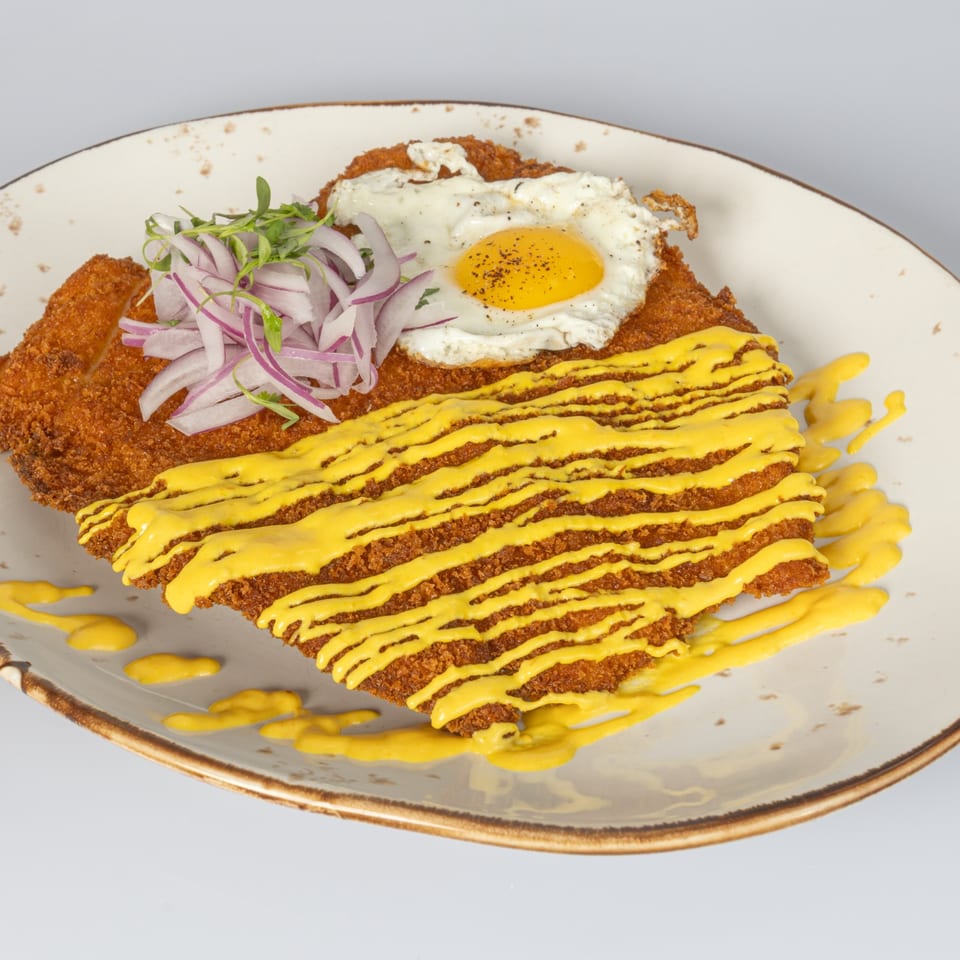 Crispy breaded cutlet topped with yellow sauce, a fried egg, sliced red onions, and microgreens on a ceramic plate.