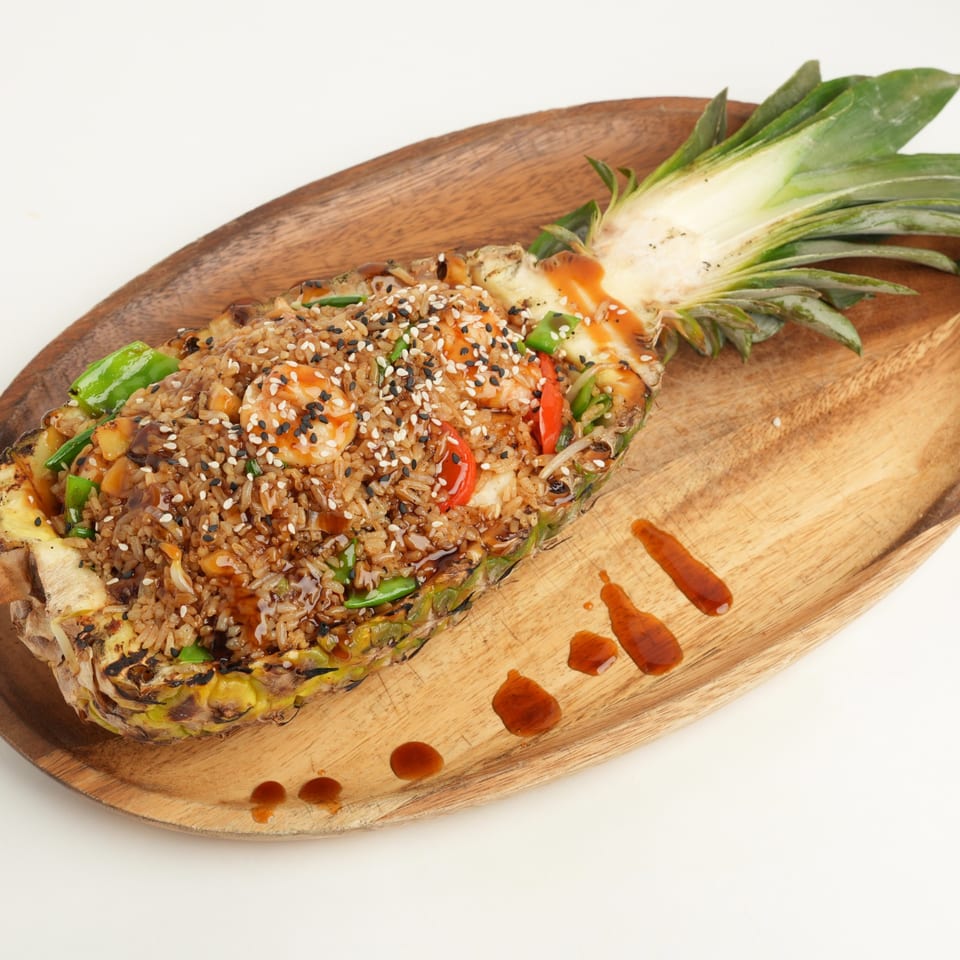 Pineapple half filled with sesame-topped fried rice, shrimp, vegetables, and drizzled sauce on a wooden serving board.