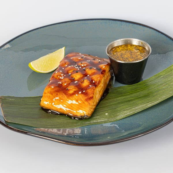 Glazed salmon with sesame seeds, a lime wedge, and a side of sauce on a banana leaf.