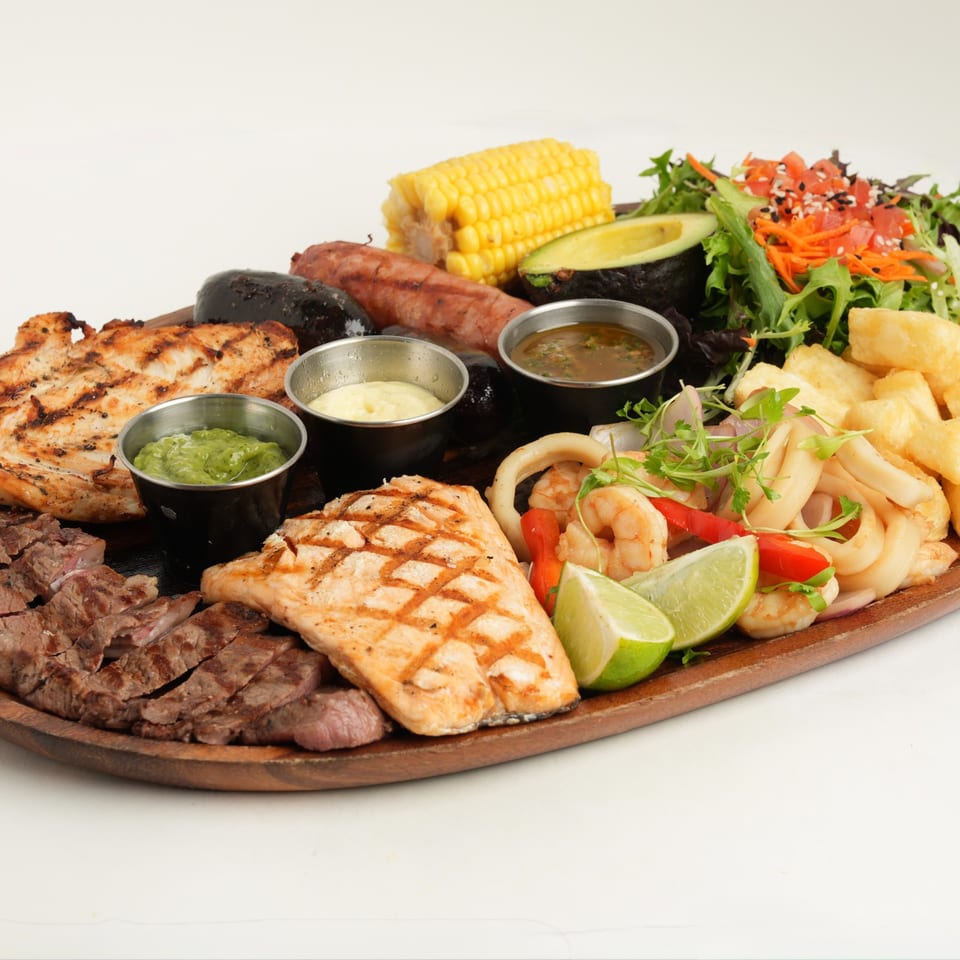 Wooden platter of grilled meats, chicken, shrimp, corn, avocado, salad, yucca, lime, and dipping sauces.